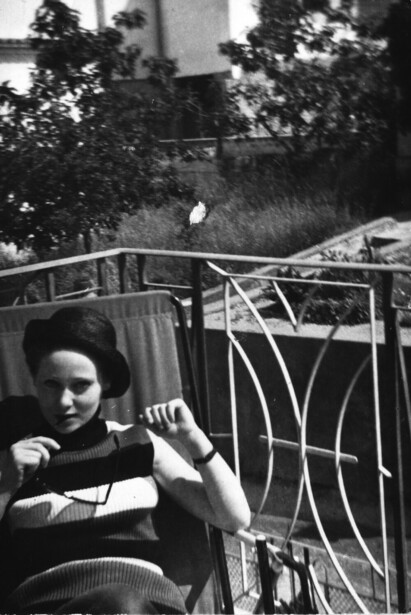 Person seated on a balcony railing, leaning against decorative metal railings with foliage visible in soft-focus background.