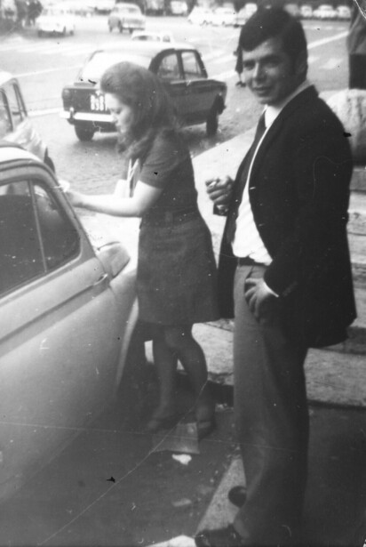 Two people standing beside a vintage car in a parking lot, black and white photograph.