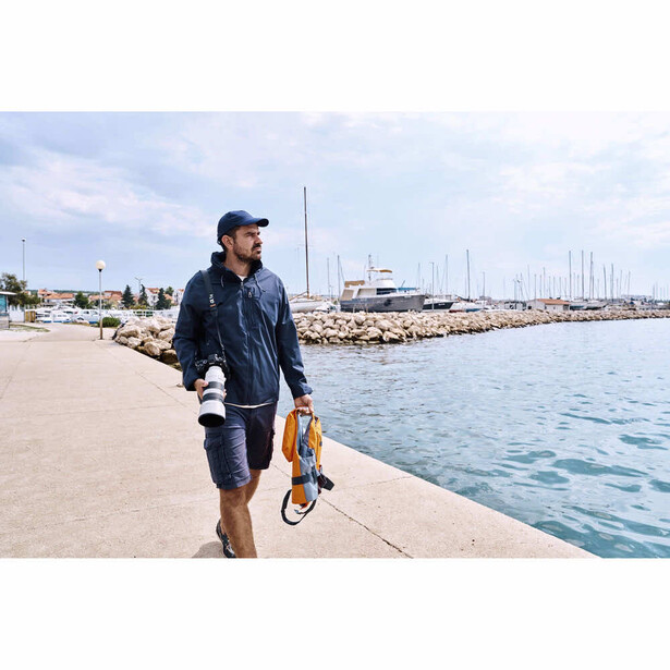 Man in dark jacket walking along a marina pier carrying skateboard shoes.