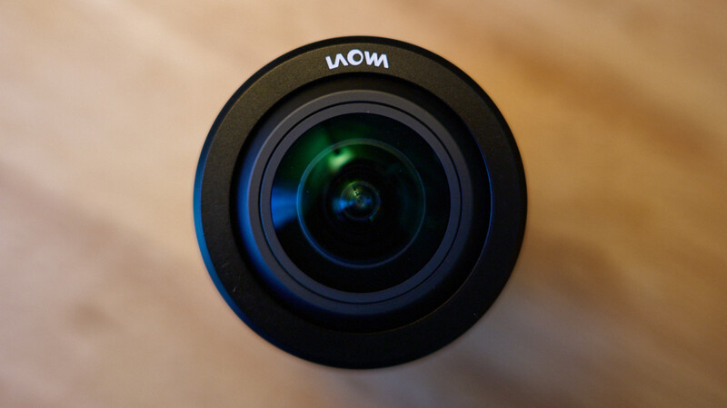 Close-up of a camera lens with green glass elements and blue ring accent against a blurred wooden background.