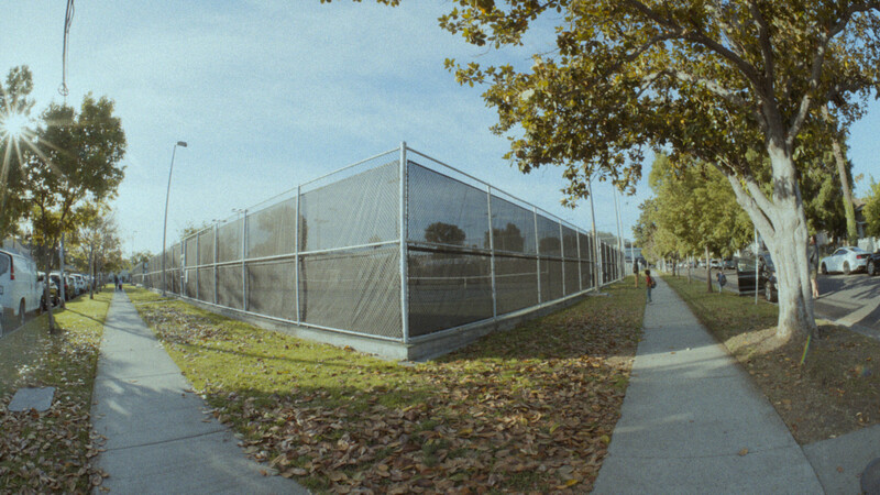 Modern greenhouse with metal frame and mesh panels in a public park setting surrounded by trees and pathways.
