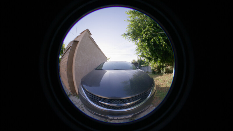 Fisheye lens view of a dark sedan parked beside a tent and trees in a rural setting.
