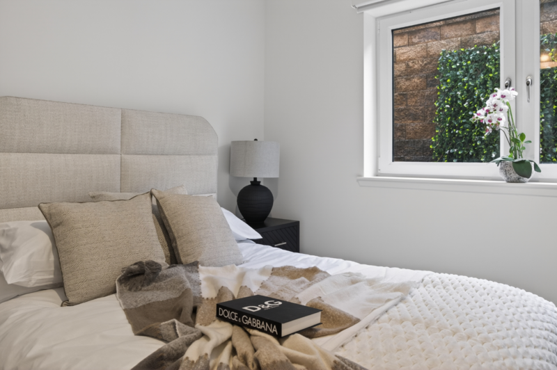 Minimalist bedroom interior with neutral bedding, decorative pillows, and a black accent object on the nightstand.