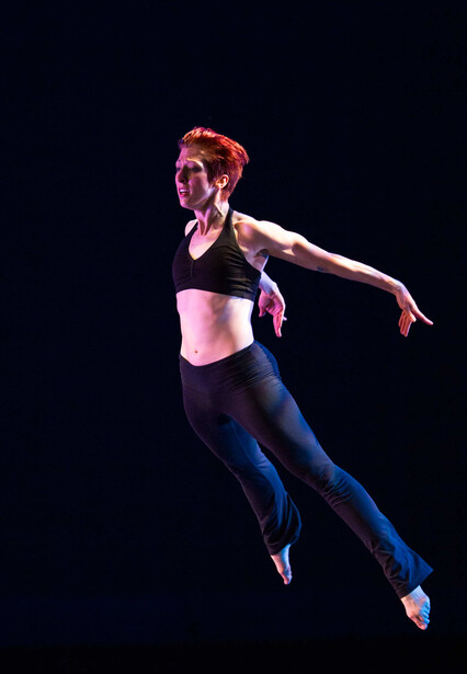 Female athlete mid-jump against black background, arms extended in dynamic pose.