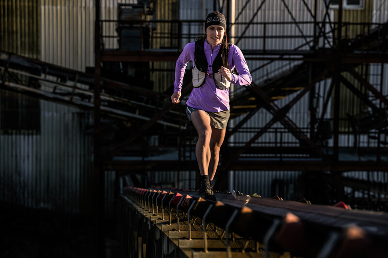 Runner in purple jacket sprinting across an industrial metal bridge with dramatic side lighting.