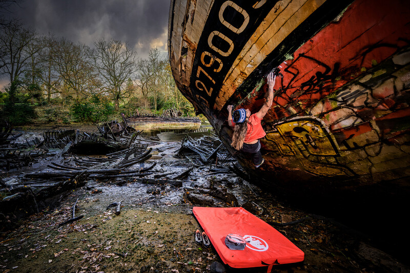 Kayaker in red jacket paddling toward a large rusty shipwreck partially submerged in a shallow river.