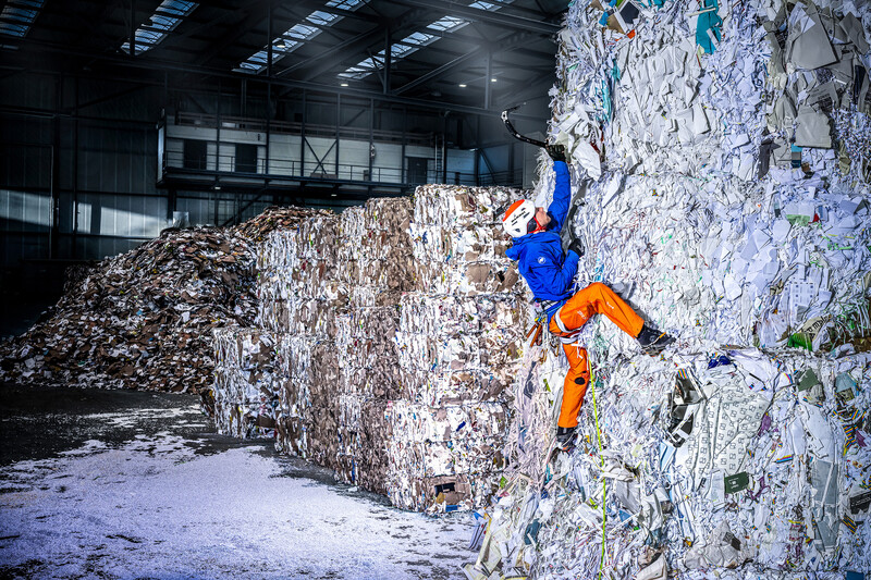 Climber in blue and orange gear ascending a steep wall of compressed recycled paper in an industrial warehouse.