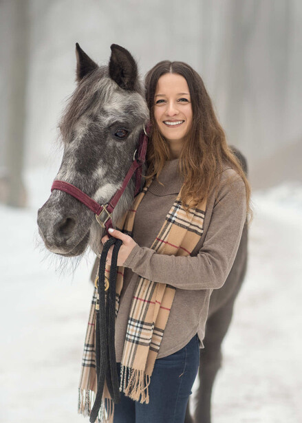 Young woman with long dark hair smiling while holding a gray horse wearing a pink halter.