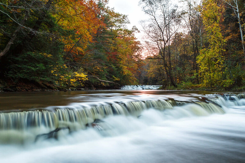 Cascading waterfall flowing through a forested river lined with autumn-colored trees.