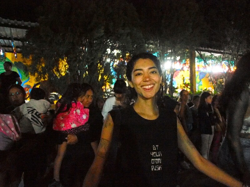 Woman smiling at camera in crowded nighttime outdoor event with colorful illuminated decorations