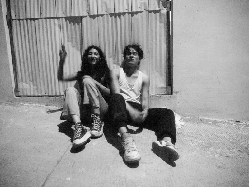 Two people sitting on concrete ground against corrugated metal wall in black and white
