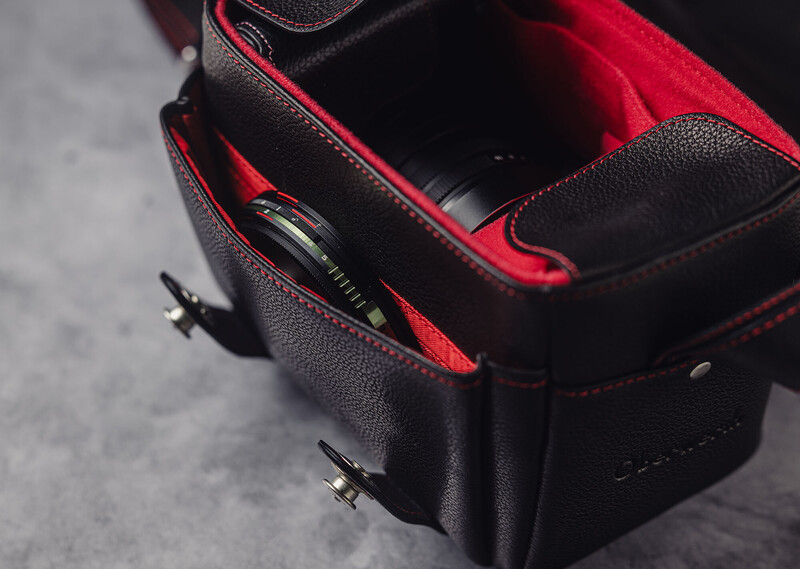 Black leather camera bag with red interior and metal clasps, photographed from above on gray surface