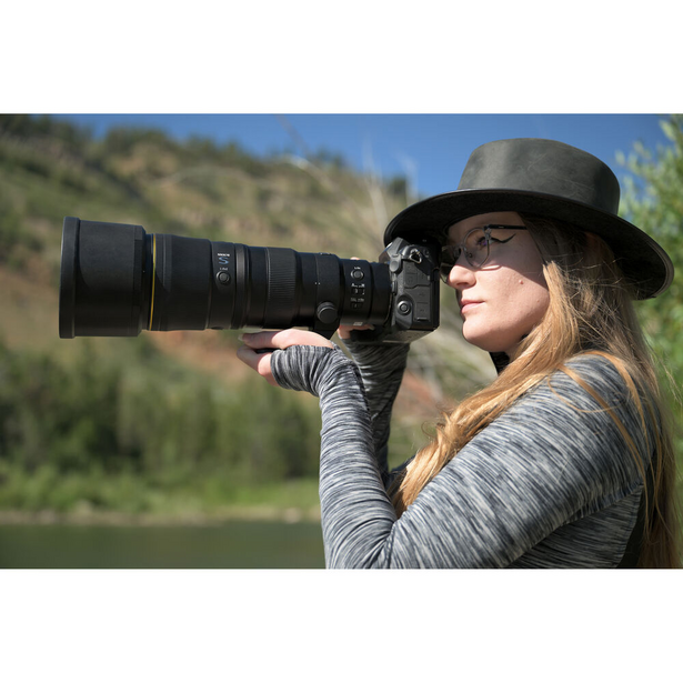 Photographer with long hair wearing a black fedora and gray jacket holds a telephoto lens camera up to her eye