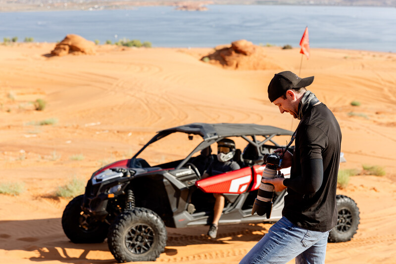 Rex Jones checking his camera on the sand dunes at Sand Hollow State Park in southern Utah, with a Can-Am side-by-side and Sand Hollow Reservoir in the background.