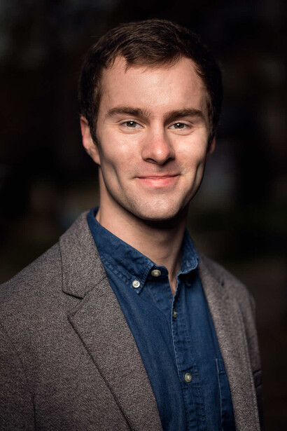 Professional headshot of a man in denim shirt and gray cardigan against dark background