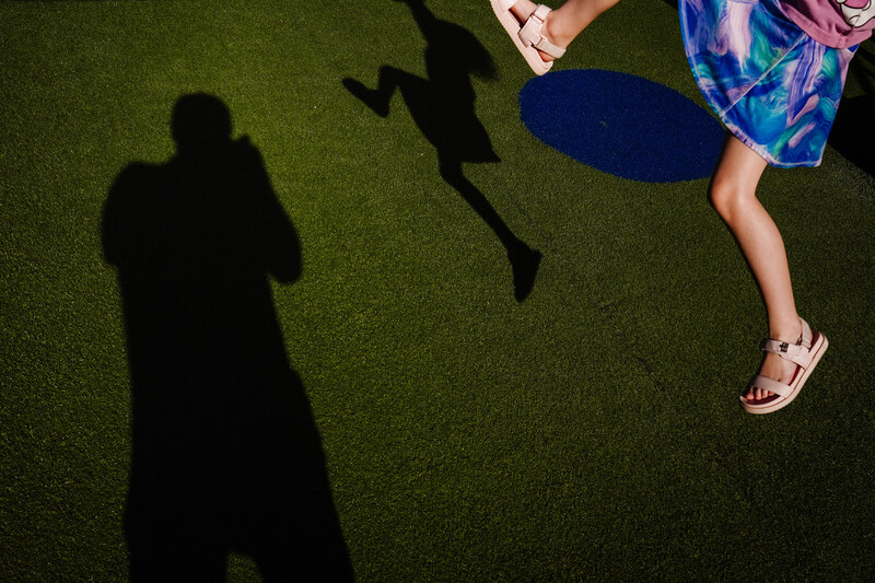 Overhead view of child's leg and shoe mid-jump on artificial grass with shadows of photographer and other figures