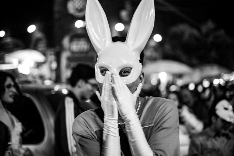 Person wearing white rabbit mask at nighttime event, hands raised to face in black and white photography