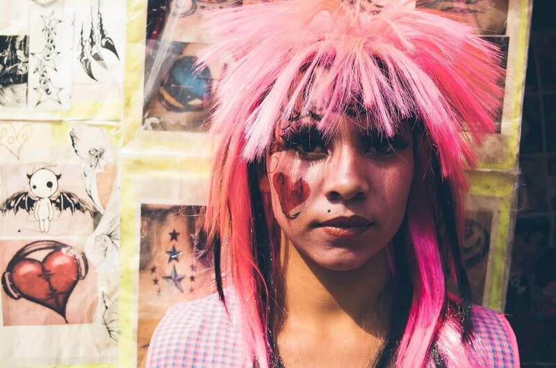 Portrait of person with bright pink wig and heart-shaped face paint against collaged wall backdrop
