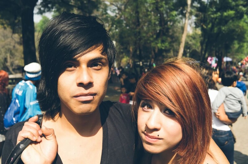 Two young people posing together outdoors in a park setting with a crowd visible in the background