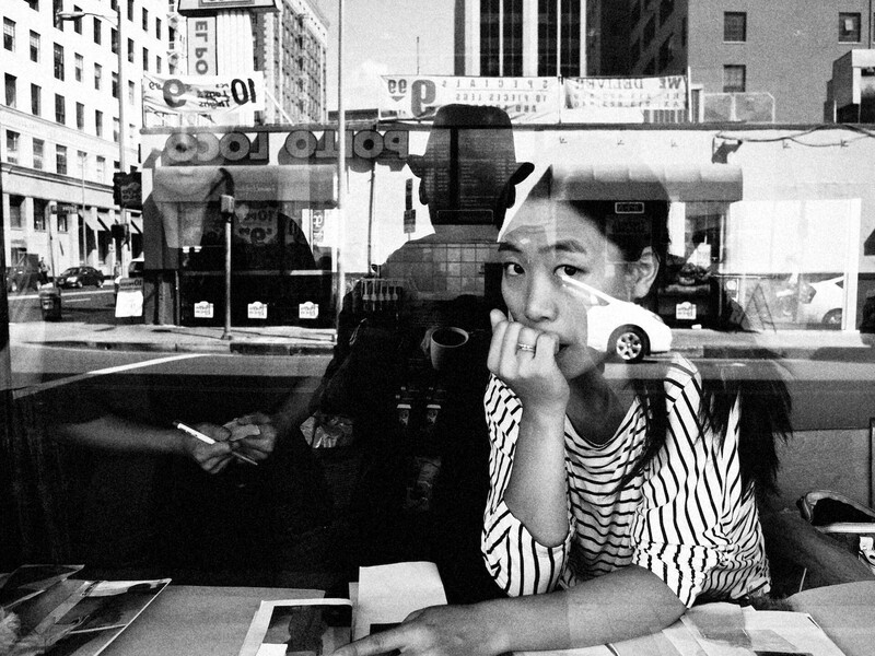 Woman in striped shirt resting chin on hand at desk, photographed through window with city street and buildings reflected behind