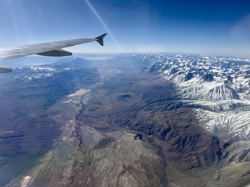 Aerial view from airplane window showing snow-capped mountains and desert valleys below