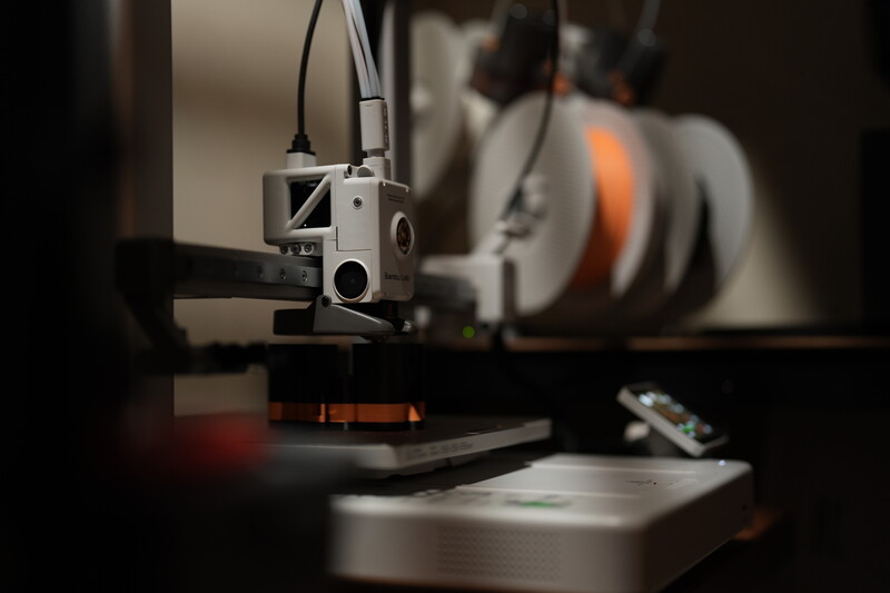 3D printer extruder head positioned over a print bed with filament spool visible in background