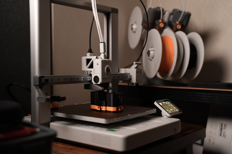 3D printer with spools of orange and white filament mounted on wall storage rack