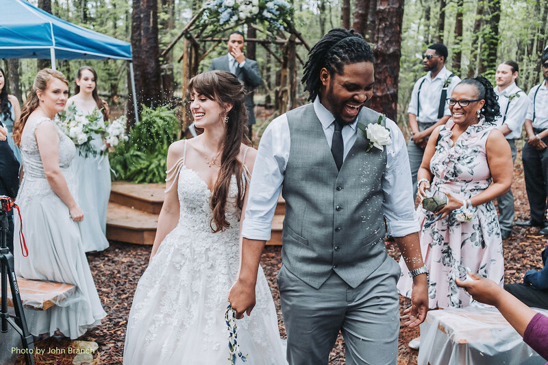 Newlyweds walking together at outdoor forest wedding ceremony with guests and blue tent visible