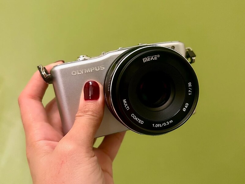 Hand holding silver Olympus mirrorless camera with black Meike lens against yellow-green background