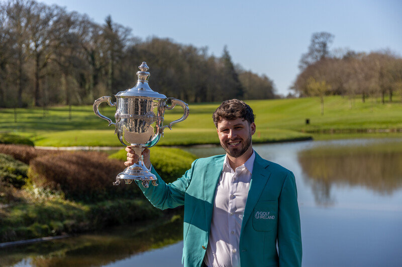 Man in teal blazer holding ornate silver trophy in park setting