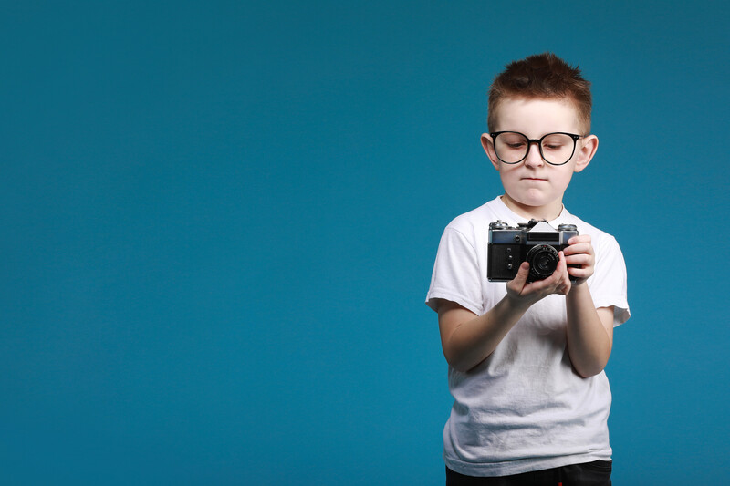 Young boy wearing glasses holding a vintage film camera against blue background