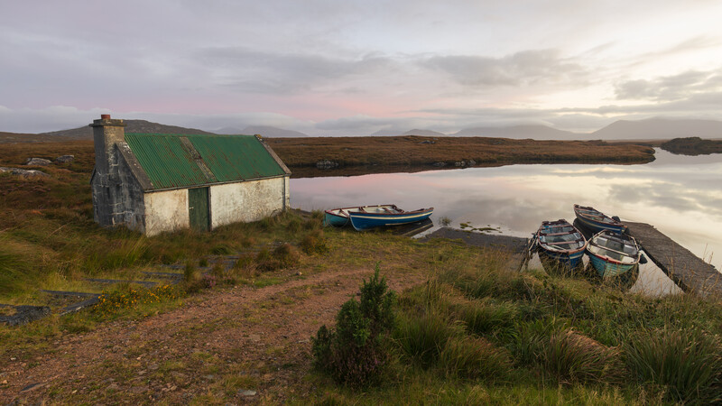 Abandoned cottage with green roof beside moored boats on calm water at dusk