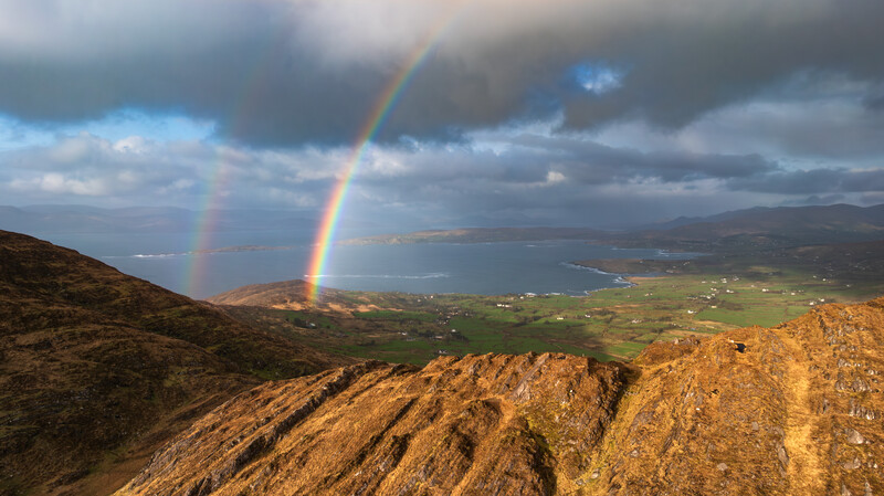Double rainbow arcing over a valley landscape with mountains and a coastal bay