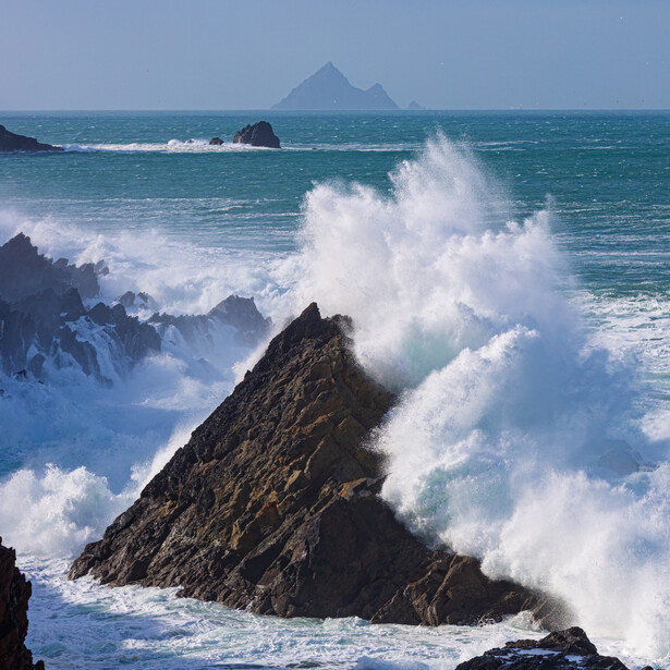 Dramatic ocean waves crashing against dark rocky outcrops with a distant mountain peak visible on the horizon