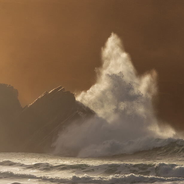 Massive ocean wave crashing against rocky coastline with dramatic backlighting