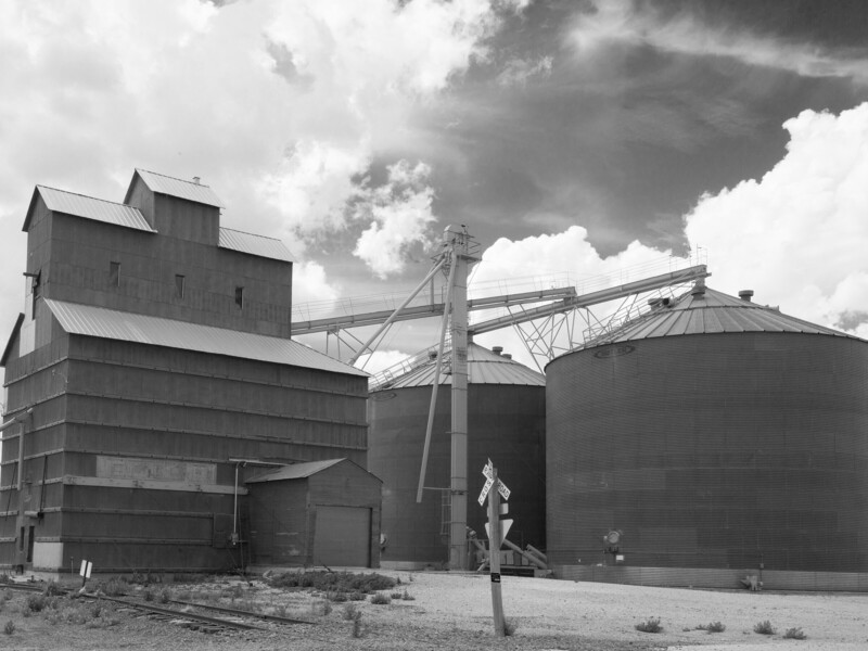 Black and white photograph of agricultural grain elevator and storage silo complex under dramatic cloudy sky