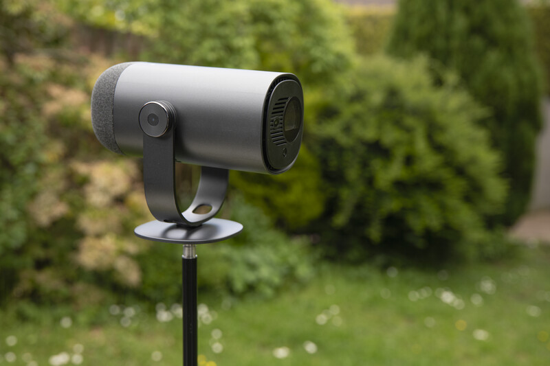 Gray cylindrical outdoor camera mounted on black stand in garden setting