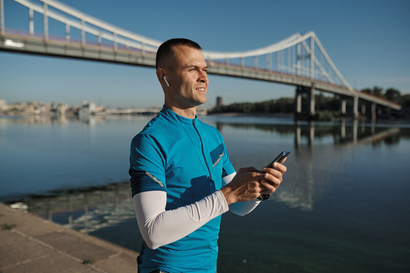 Man in blue athletic shirt holding smartphone by waterfront with suspension bridge