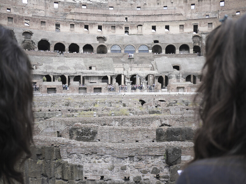 Two figures viewing ancient Roman amphitheater arena from upper seating area