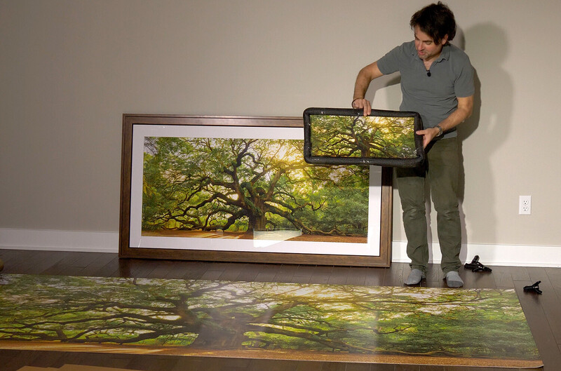 Man adjusting a tablet displaying a tree image alongside framed landscape prints on white gallery wall