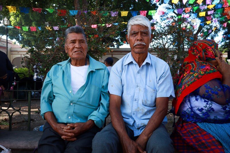 Two men seated together outdoors beneath a tree decorated with colorful paper banners