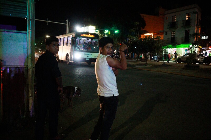 Young man in white sleeveless shirt on nighttime city street with bus and pedestrians