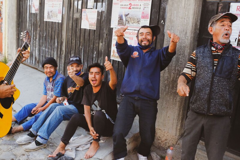 Five musicians celebrating outdoors against a weathered concrete wall, with one holding an acoustic guitar