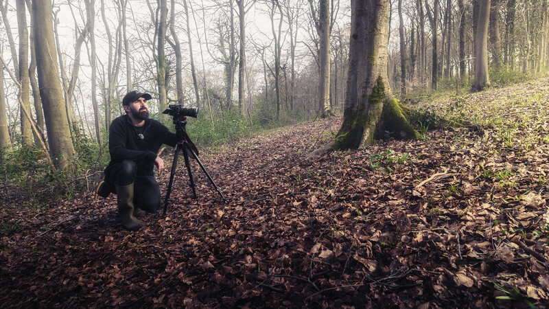 Photographer kneeling with telephoto lens on tripod in autumn forest with fallen leaves