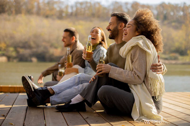 Four friends sitting on a wooden dock by a lake, holding beer bottles and laughing together