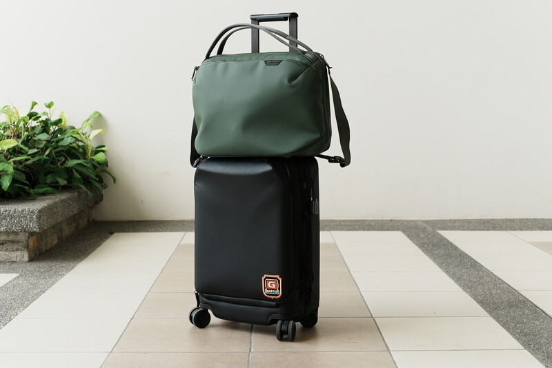 Black rolling luggage with green canvas travel bag stacked on top, displayed on tiled floor
