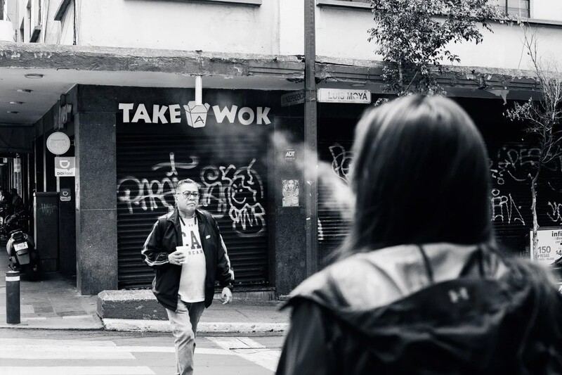 Black and white street photography of a person walking past a storefront with "TAKE WOK" signage and graffiti