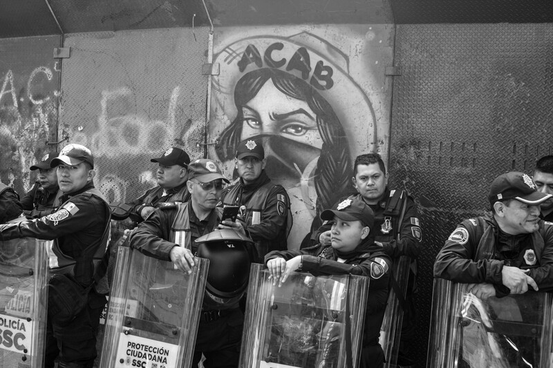 Police in riot gear standing before mural with portrait of woman wearing mask