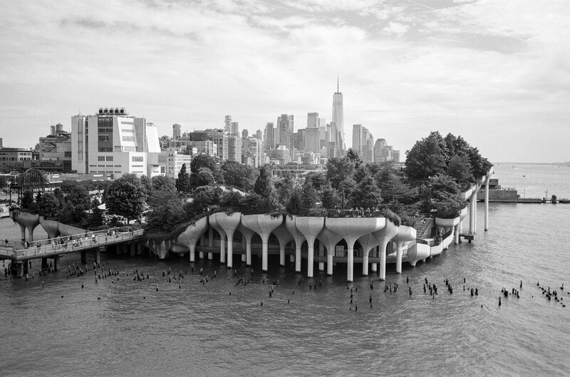 Black and white photograph of a ribbed concrete waterfront structure with Manhattan skyline in background