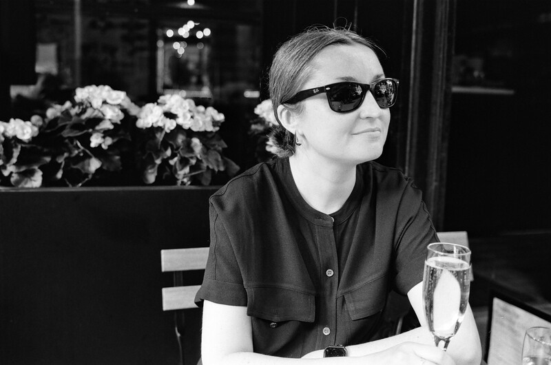 Person wearing sunglasses and black shirt seated at outdoor café table in black and white portrait